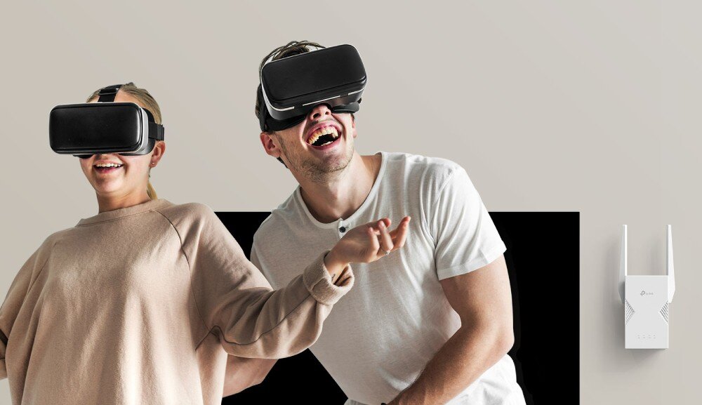 Dwie osoby noszą gogle VR, uśmiechają się i gestykulują. Obok na ścianie znajduje się wzmacniacz sieci TP-LINK z widocznym logo producenta.
