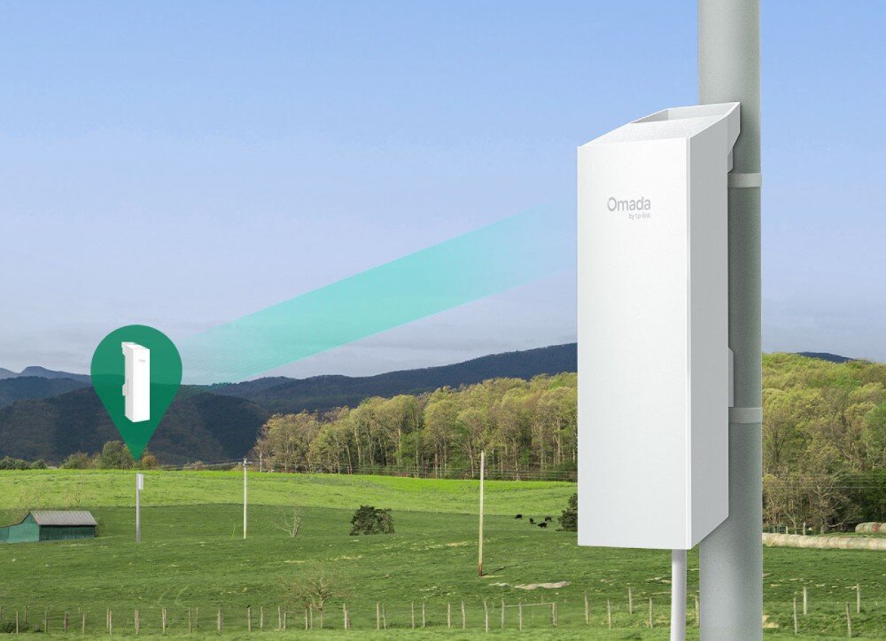 Most bezprzewodowy TP-LINK Omada EAP100-Bridge Kit Białe, podłużne urządzenie zamontowane na szarym słupie na tle zielonych pól i wzgórz, wysyłające niebieską wiązkę sygnału w stronę drugiego, oddalonego punktu