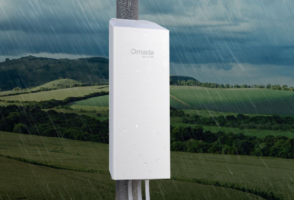 Most bezprzewodowy TP-LINK Omada EAP100-Bridge Kit Białe urządzenie w obudowie odpornej na warunki atmosferyczne, pokryte kroplami wody, zamontowane na słupie podczas deszczowej pogody na tle krajobrazu