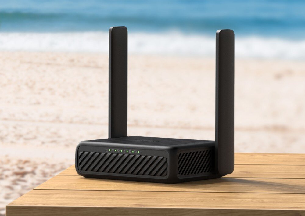 Czarny router z dwoma pionowymi antenami stoi na drewnianym stole, w tle widać plażę i morze. Na przednim panelu routera znajdują się diody sygnalizacyjne.