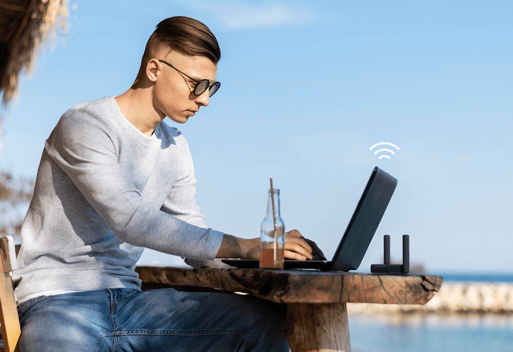 Osoba siedzi przy drewnianym stole na zewnątrz, używając laptopa. Obok laptopa znajduje się router z dwiema antenami, a w tle widać morze i niebo.