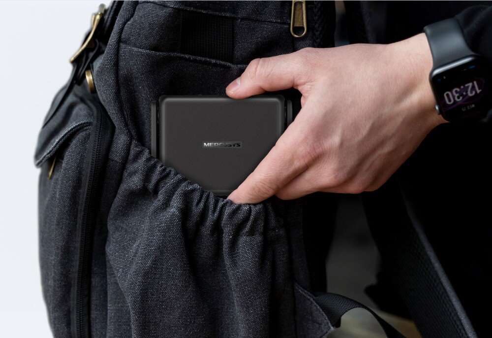 Osoba wkłada czarne urządzenie z logo \'MERCUSYS\' do kieszeni plecaka. Na nadgarstku widoczny jest zegarek z wyświetlaczem pokazującym godzinę 12:30.