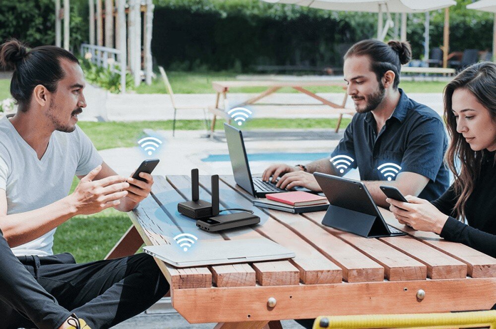 Na drewnianym stole znajduje się router z dwiema antenami, wokół którego siedzą trzy osoby korzystające z urządzeń mobilnych. Nad urządzeniami widoczne są symbole sygnału Wi-Fi.