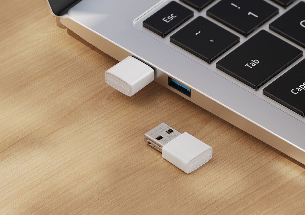 Karta sieciowa MERCUSYS MA60XNB Biała karta sieciowa o niewielkich rozmiarach wpięta do bocznego portu USB srebrnego laptopa obok klawiszy Esc i Tab oraz druga taka sama karta leżąca obok na jasnym drewnianym blacie