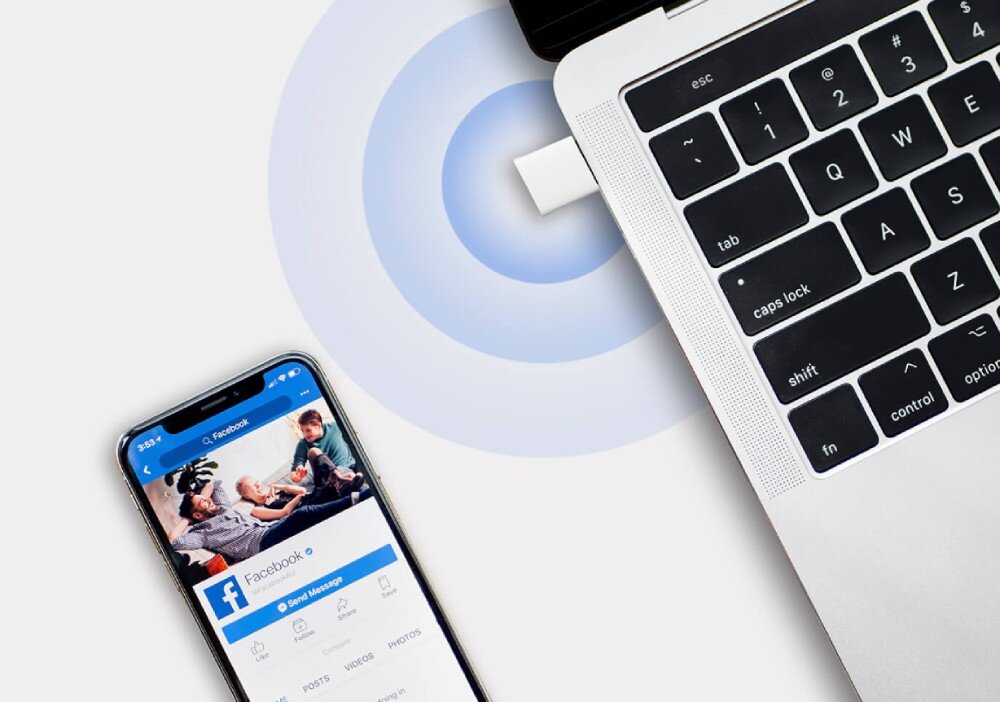 Karta sieciowa MERCUSYS MA60XNB Widok z góry na róg srebrnego laptopa z wpiętą kartą sieciową która emituje niebieskie fale sygnału w stronę leżącego obok smartfona z uruchomioną aplikacją społecznościową