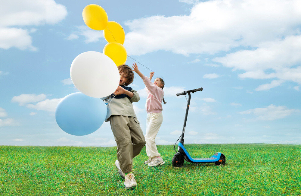 Hulajnoga elektryczna SEGWAY Ninebot C2 Lite 9km 130W 7 cali Czarno-niebieski łąka trawnik niebo dwoje dzieci bawi się balonami obok stoi hulajnoga przygoda bezpieczeństwo komfort wygoda radość zabawa