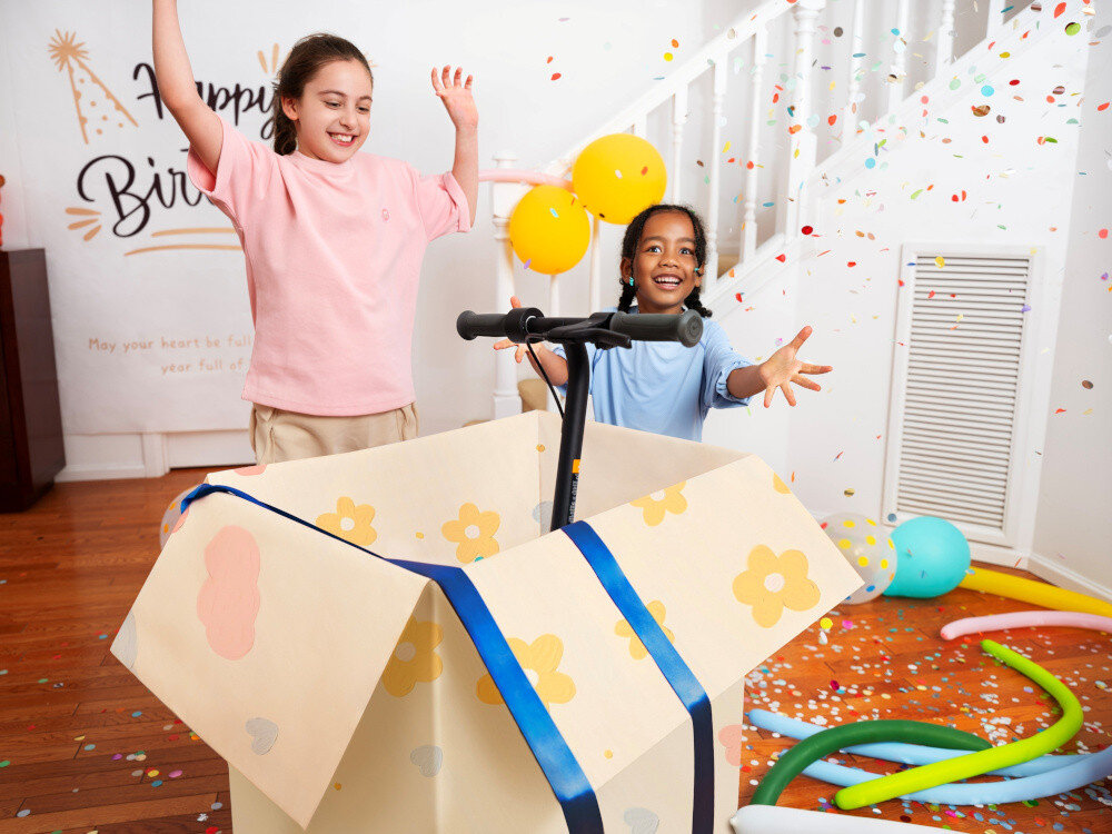 Hulajnoga elektryczna SEGWAY Ninebot C2 Lite 9km 130W 7 cali Czarno-niebieski pomieszczenie dwójka dzieci otwiera karton z prezentem w kartonie hulajnoga w tle schody konfetti dla dzieci w wieku od 7 do 10 lat zalecany wzrost dziecka 115–145 cm lekka konstrukcja intuicyjna obsługa odpowiednio dobrane parametry techniczne bezpieczeństwo radość jazdy