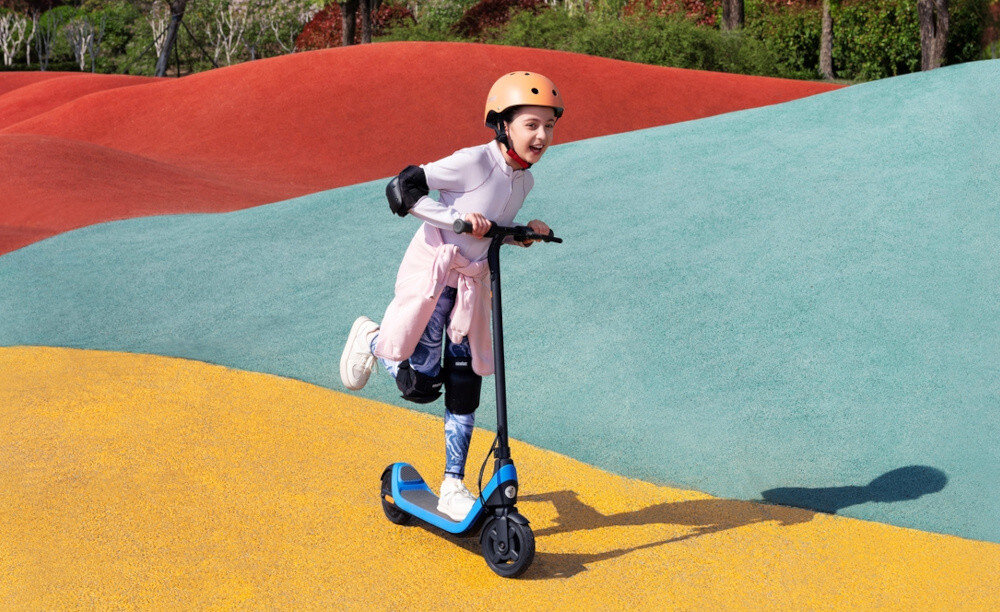 Hulajnoga elektryczna SEGWAY Ninebot C2 Lite 9km 130W 7 cali Czarno-niebieski skatepark kolorowa droga dziecko jedzie na hulajnodze w tle drzewa rośliny waga 7,8 kg wykonana ze stali obciążenie do 50 kg dla dzieci w wieku od 7 do 10 lat stabilność i bezpieczeństwo