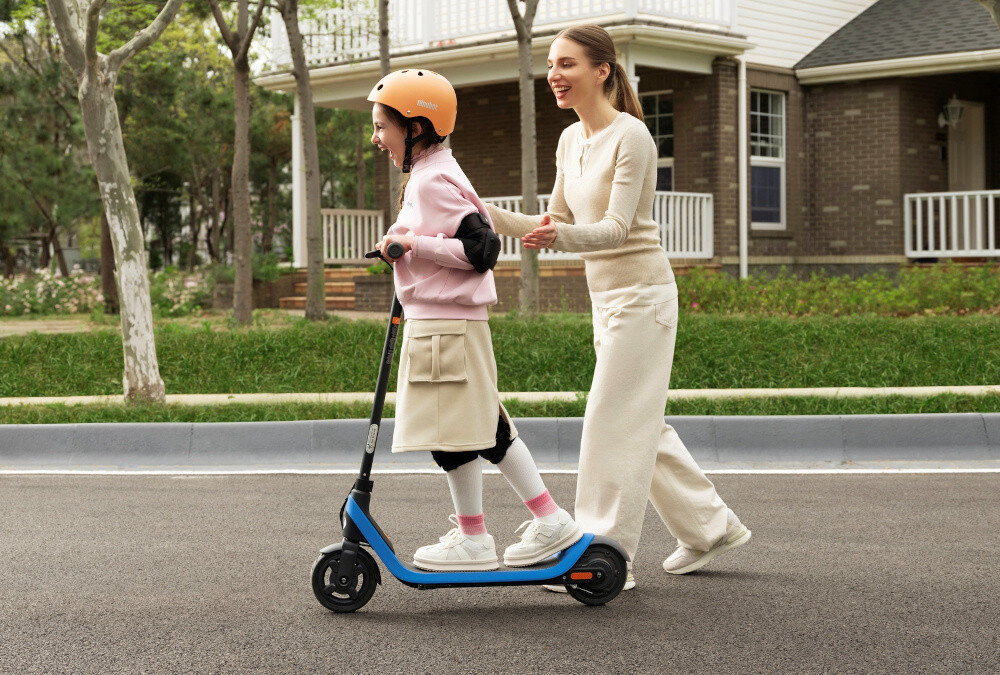 Hulajnoga elektryczna SEGWAY Ninebot C2 Lite 9km 130W 7 cali Czarno-niebieski droga miasto dziecko jedzie na hulajnodze obok dziecka idzie dorosła osoba w tle dom trawnik krzewy klasa wodoodporności IPX4 ochrona przed zachlapaniem i deszczem certyfikaty RoHS i REACH brak substancji niebezpiecznych bezpieczeństwo dla zdrowia dziecka trwałość konstrukcji bezpieczeństwo użytkowania