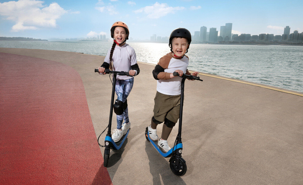 Hulajnoga elektryczna SEGWAY Ninebot C2 Lite 9km 130W 7 cali Czarno-niebieski chodnik droga dwoje dzieci jedzie na dwóch hulajnogach uśmiechają się w tle budynki miasta woda rzeka 7-calowe pełne opony bezdętkowe stabilna i bezpieczna jazda bez ryzyka nagłej utraty powietrza opony skutecznie amortyzują drobne nierówności nawierzchni płynna i stabilna jazda