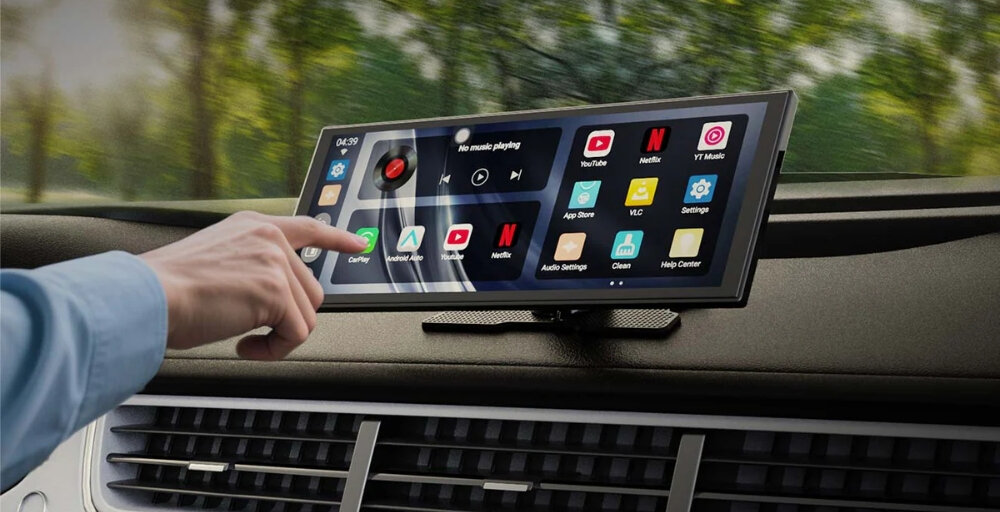 Tablet zamontowany na desce rozdzielczej samochodu wyświetla interfejs z ikonami aplikacji, takimi jak YouTube, Netflix i CarPlay. Osoba dotyka ekranu, na którym widoczny jest także zegar i pasek odtwarzania muzyki.