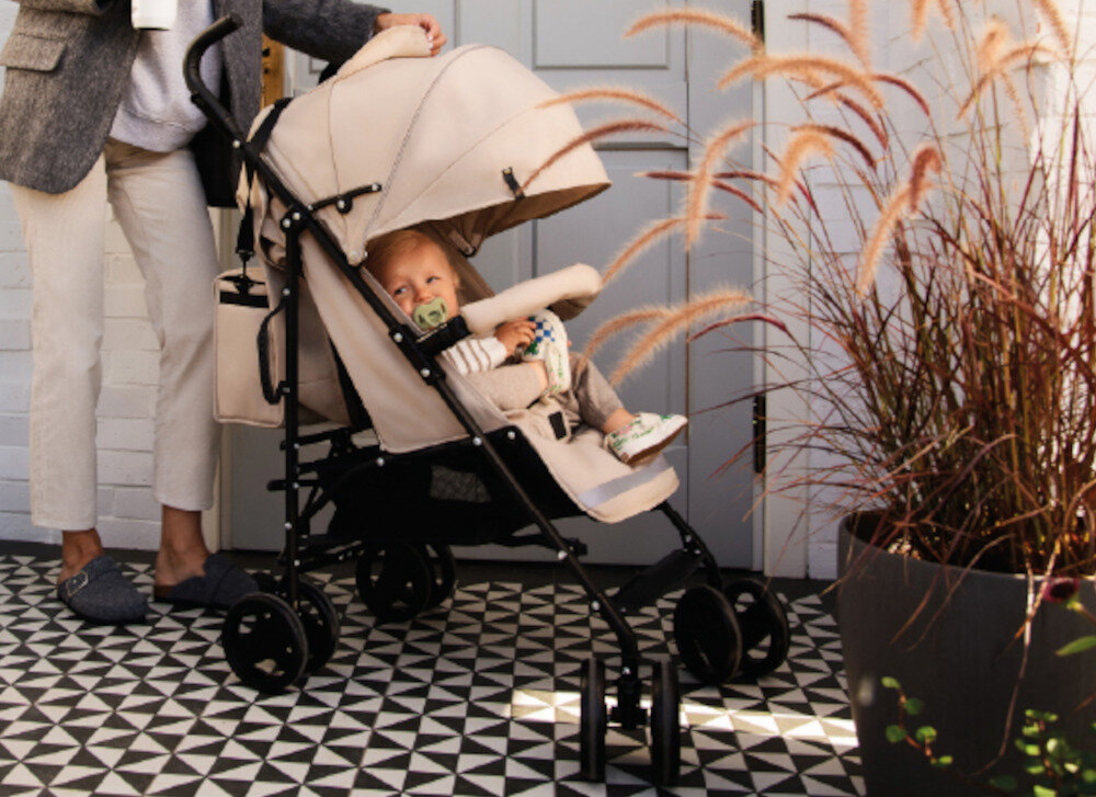 Beżowy wózek spacerowy z dzieckiem siedzącym w środku, obok stoi osoba trzymająca kubek. W tle widoczne są drzwi i rośliny w donicy.