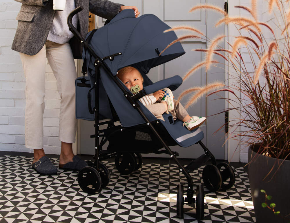 Na zdjęciu znajduje się szary wózek spacerowy z dzieckiem siedzącym w środku. W tle widać osobę stojącą obok wózka oraz rośliny w donicy na podłodze z geometrycznym wzorem.