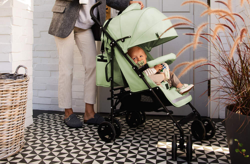 Na zdjęciu znajduje się zielony wózek spacerowy z dzieckiem w środku. W tle widać osobę dorosłą oraz elementy dekoracyjne, takie jak kosz wiklinowy i rośliny.