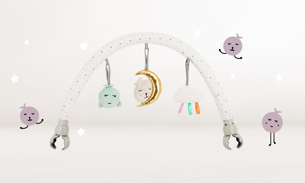 Pałąk z zabawkami w kształcie łuku, z trzema wiszącymi pluszowymi zabawkami: zieloną rybką, księżycem i chmurką. Tło jest jasne z narysowanymi uśmiechniętymi buźkami i gwiazdkami.