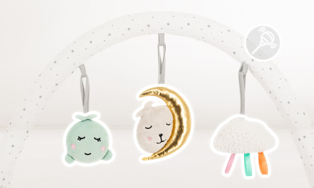 Na obrazie widoczny jest biały pałąk z szarymi gwiazdkami, na którym zawieszone są trzy pluszowe zabawki: zielona postać, księżyc z twarzą oraz chmurka z kolorowymi tasiemkami. W prawym górnym rogu znajduje się symbol dźwięku.