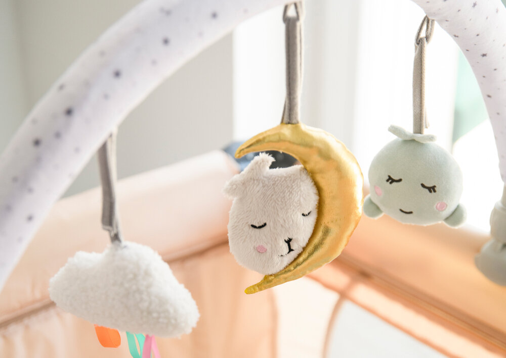 Na zdjęciu widać pałąk z zabawkami, na którym wiszą trzy pluszowe zawieszki: chmurka, księżyc z twarzą oraz zielona kulka z uśmiechem. Pałąk jest biały z drobnymi czarnymi kropkami.