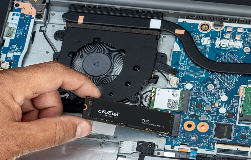 Dysk CRUCIAL T500 Ręka trzymająca cienki dysk SSD podczas montażu wewnątrz laptopa obok układów i wentylatora, aplikacja, konfiguracja, instalacja
