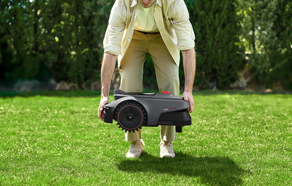 Osoba trzyma czarny robot koszący na trawniku. Na robocie widoczne są napisy \'MOVA\' i \'ViAX\'.