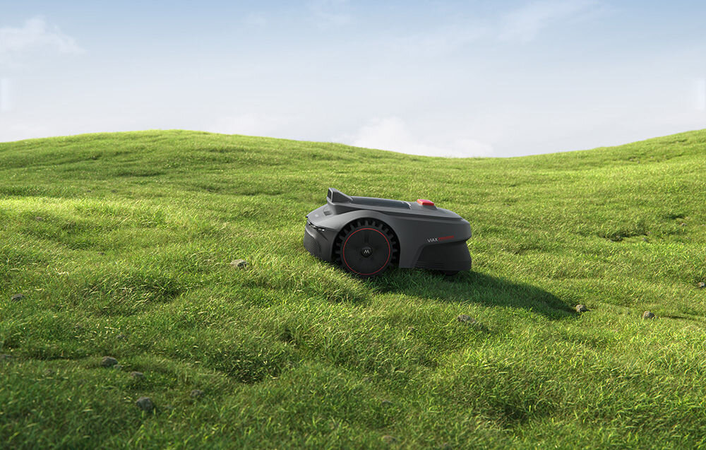 Na zielonej trawie znajduje się ciemnoszary robot koszący z czerwonymi akcentami. W tle widać niebo z lekkimi chmurami.