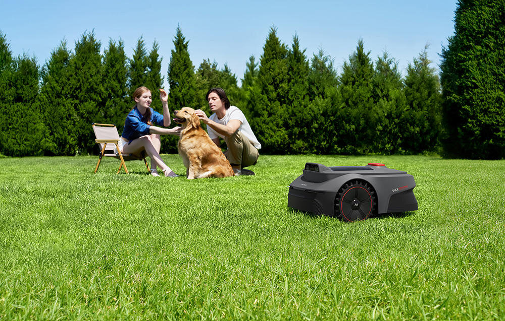 Na trawniku znajduje się czarny robot koszący z czerwonymi akcentami. W tle widoczne są dwie osoby siedzące na trawie z psem, a za nimi rząd drzew iglastych.