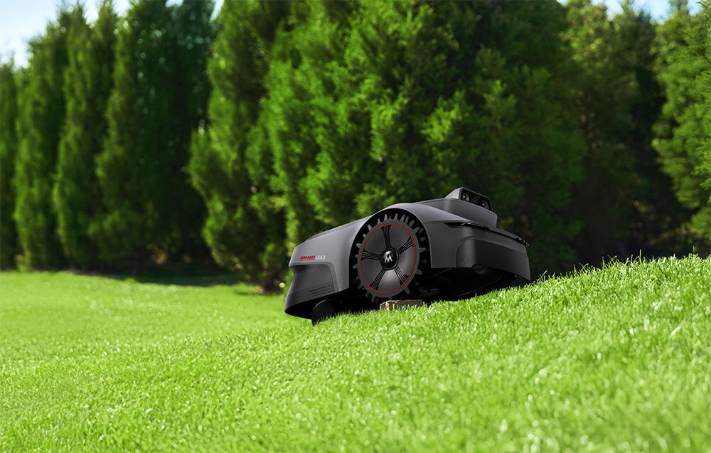 Czarny robot koszący porusza się po trawniku, w tle widoczne są drzewa iglaste. Na robocie znajduje się napis \'MOVA ViAX 300\'.