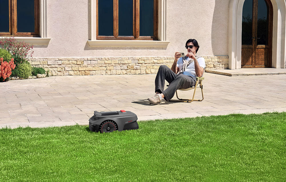 Na trawniku znajduje się robot koszący, a w tle siedzi osoba na krześle ogrodowym, trzymająca telefon. Budynek z dużymi oknami i kamienną elewacją widoczny jest w tle.