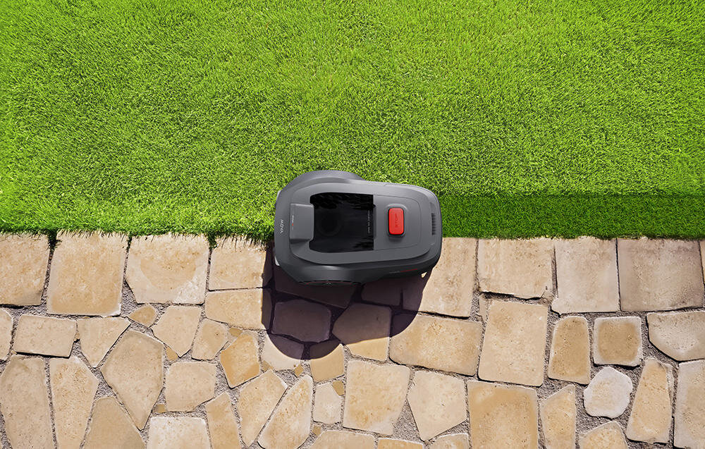 Robot koszący porusza się po granicy trawnika i kamiennej ścieżki. Urządzenie jest ciemnoszare z czerwonym przyciskiem na górze.