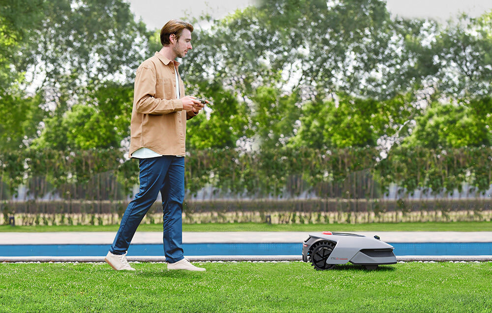 Robot koszący DREAME A1 PRO 1000 ogród trawnik robot kosi trawę za robotem idzie osoba z telefonem w dłoni zadbany trawnik prosta obsługa bezpieczeństwo wygoda