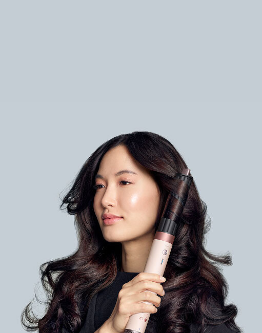 Modelka z ciemnymi włosami kręci loki używając multistylera Dyson Airwrap Coanda2x z końcówką cylindryczną. Wersja mobile.