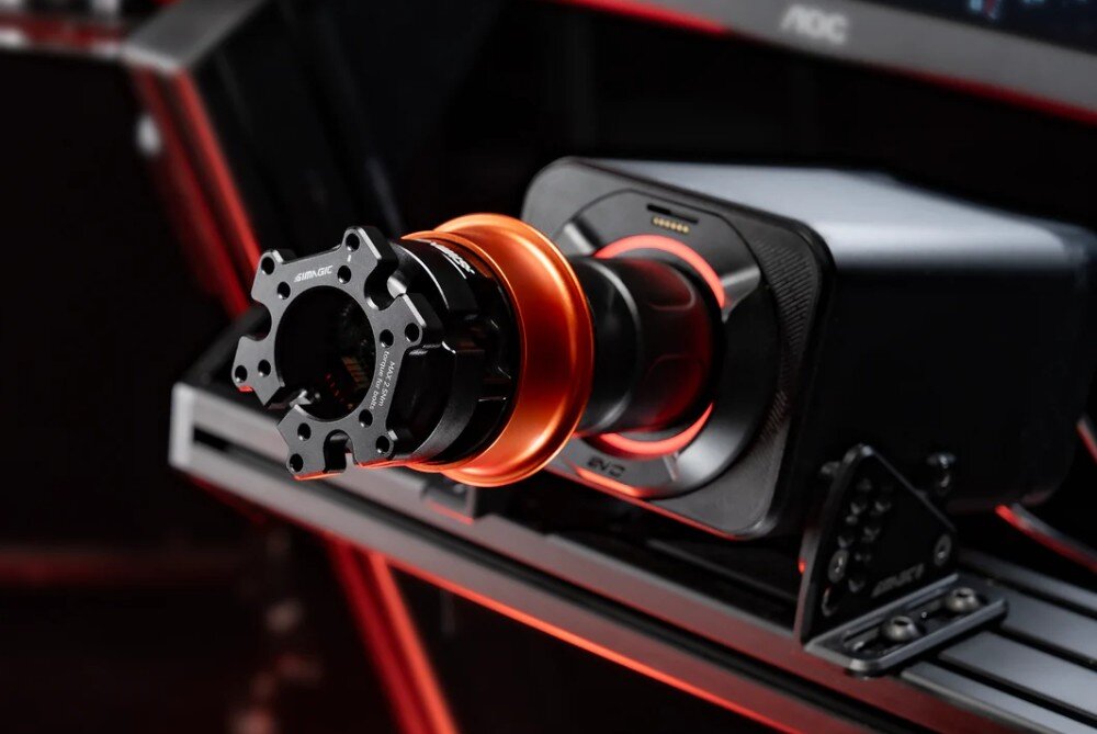 Na obrazie widoczny jest adapter SIMAGIC QR-A w kolorze czarnym z pomarańczowym elementem, zamontowany na urządzeniu przypominającym symulator kierownicy. W tle widać fragmenty konstrukcji z czerwonymi akcentami.