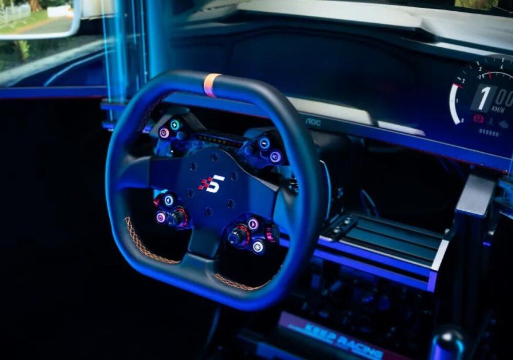 Na obrazie widoczna jest kierownica z logo \'S\' zamontowana w symulatorze jazdy. W tle znajduje się ekran z prędkościomierzem pokazującym 100 km/h.