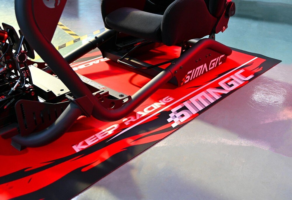 Mata pod fotel gamingowy SIMAGIC Carpet Widok od przodu na konstrukcję fotela wyścigowego stojącą na czerwonej macie z białym napisem 'KEEP RACING' i logo marki