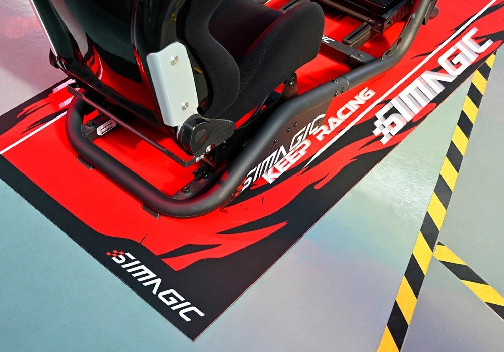 Mata pod fotel gamingowy SIMAGIC Carpet Widok z góry na konstrukcję fotela wyścigowego stojącą na czerwonej macie z widocznym logo marki w rogach i napisem 'KEEP RACING' na środku boku