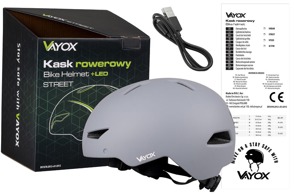 Na obrazie znajduje się szary kask rowerowy z logo VAYOX, obok którego leży kabel USB oraz pudełko z napisem \'Kask rowerowy Bike Helmet +LED STREET\'. Widoczna jest także ulotka z informacjami o produkcie i logo VAYOX z hasłem \'MOVE ON & STAY SAFE WITH\'.