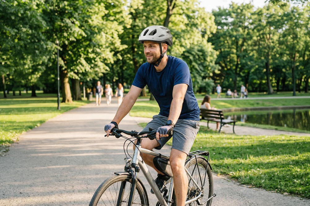 Osoba na rowerze w parku, nosząca biało-czarny kask rowerowy. W tle widoczne są drzewa, ławka i kilka osób spacerujących.
