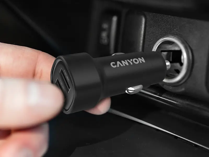 Czarna ładowarka samochodowa z dwoma portami USB-A jest wkładana do gniazda zapalniczki. Na obudowie widoczne jest logo \'CANYON\'.