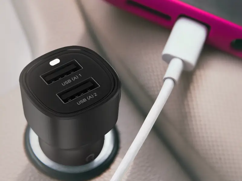 Czarna ładowarka samochodowa z dwoma portami USB oznaczonymi jako \'USB (A) 1\' i \'USB (A) 2\'. Obok ładowarki widoczny jest biały kabel podłączony do różowego urządzenia.