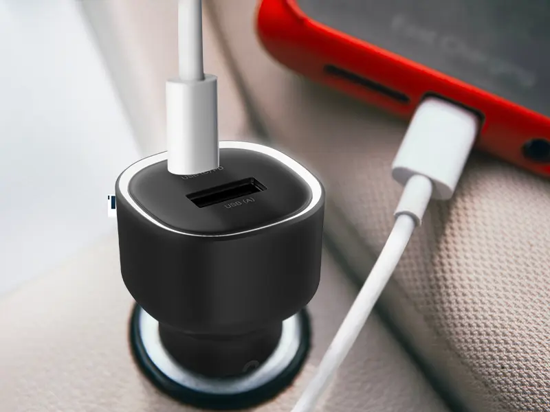 Czarna ładowarka samochodowa z dwoma portami USB, podłączona do gniazda zapalniczki. Obok znajduje się czerwony telefon z podłączonym kablem do ładowania.