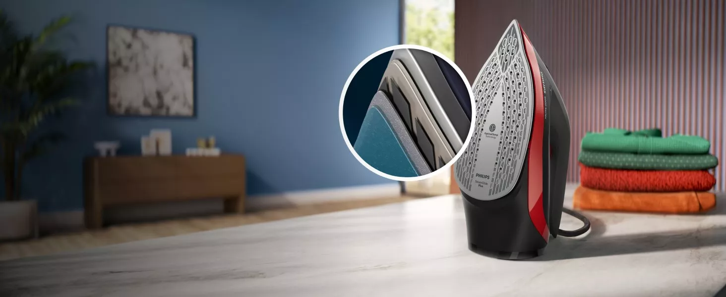 Żelazko z widoczną stopą grzewczą z napisem \'PHILIPS SteamGlide Plus\' stoi na blacie. W tle znajduje się stos złożonych ubrań oraz mebel na tle niebieskiej ściany.