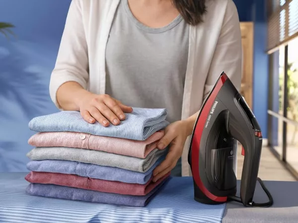 Osoba układa stos kolorowych koszul na stole, obok stoi żelazko z widocznym napisem \'Philips 6000 Series\'.