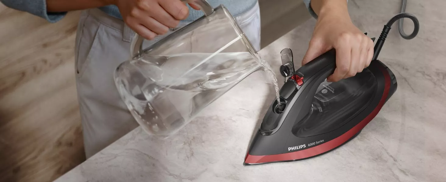Osoba nalewa wodę z dzbanka do żelazka marki PHILIPS 6000 Series, które znajduje się na blacie. Żelazko ma czarną i czerwoną obudowę z widocznym logo PHILIPS.