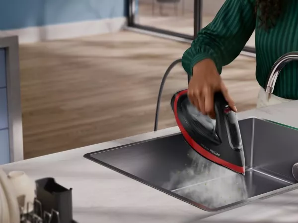 Osoba trzyma żelazko nad zlewem, z którego wydobywa się para. Na żelazku widoczne jest logo \'Philips\'.