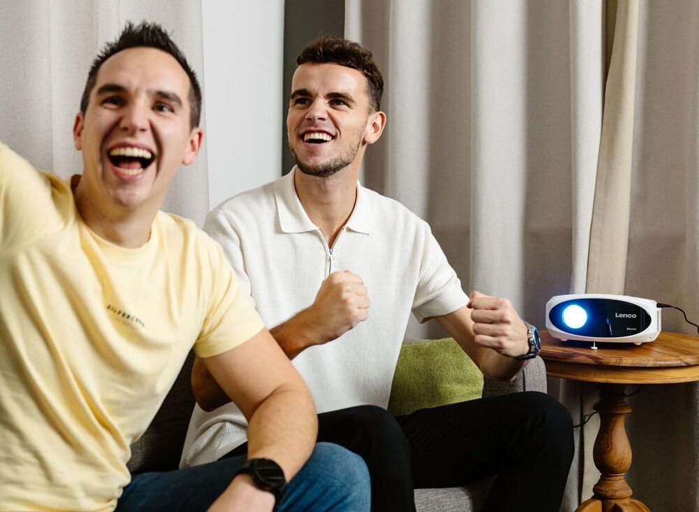 Projektor LENCO LPJ-900WH projekcja 1-7 m 510 cm 200 cali korekcja trapezowa pionowa Full HD, osoby siedzące na kanapie, projektor stojący na stoliku