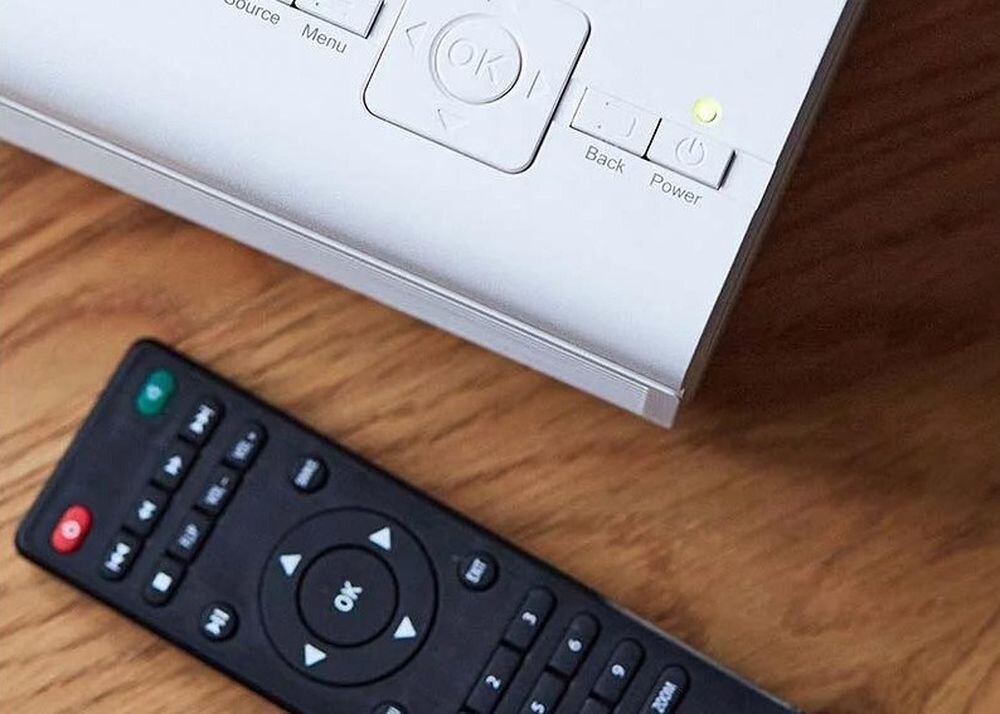 Projektor LENCO LPJ-280WH WVGA 800 x 480 2800 ANSI lumen pilot zdalnego sterowania 2x AAA zasilanie 100-240 V Typ C, kadr pojektora oraz pilota leżącego na blacie