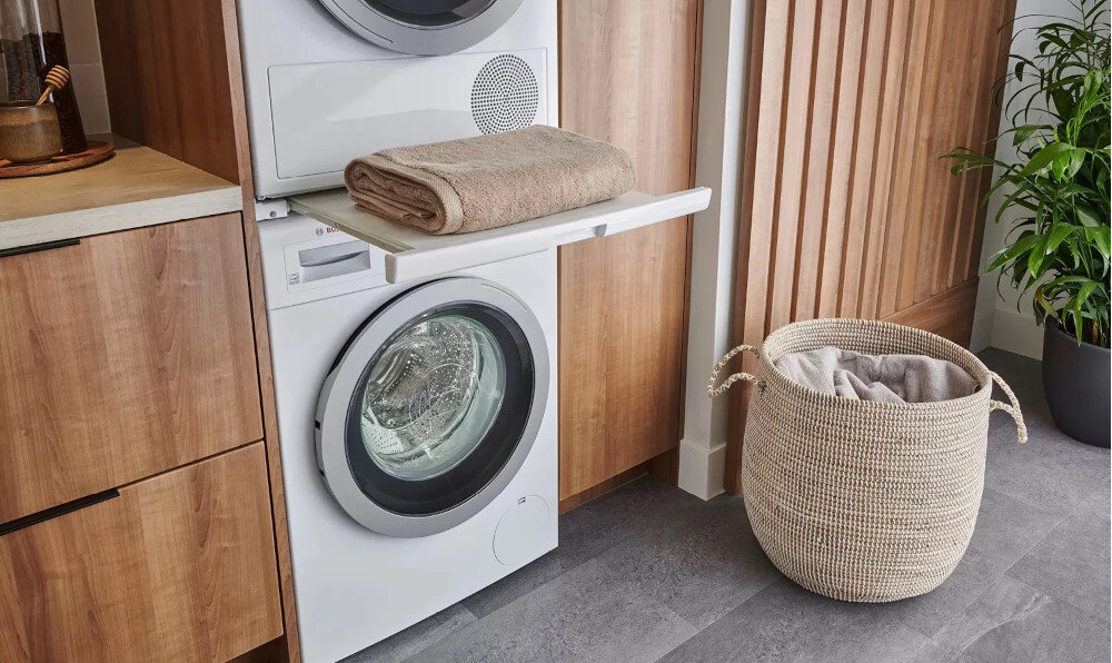 SUSZARKA BOSCH WQG244CSPL Na zdjęciu widoczna jest pralka i suszarka ustawione jedna na drugiej w zabudowie z drewnianymi elementami. Przed pralką wysunięta jest praktyczna półka złożonym ręcznikiem, ułatwiająca odkładanie prania. Obok stoi kosz na bieliznę, a z prawej strony widać zieloną roślinę doniczkową ustawienie na pralce stabilność urządzeń łącznik suszarka-pralka wygodne przenoszenie prania bezpieczne użytkowanie szybkie pranie i suszenie