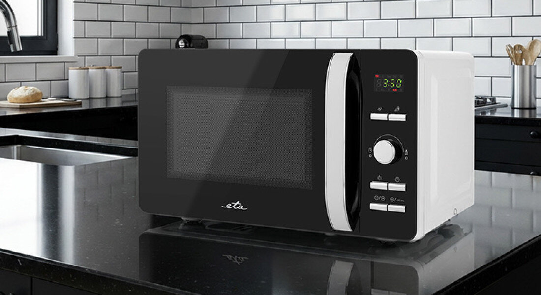 Kuchenka mikrofalowa ETA Gavento ETA120990000 Pierwsze zdjęcie przedstawia wolnostojącą kuchenkę mikrofalową marki ETA ustawioną na czarnym, połyskującym blacie w kuchni z białymi płytkami i czarną armaturą. Urządzenie ma czarny front z wyświetlaczem LED oraz pokrętłami po prawej stronie, a jasne światło z okna podkreśla jego połyskujące wykończenie