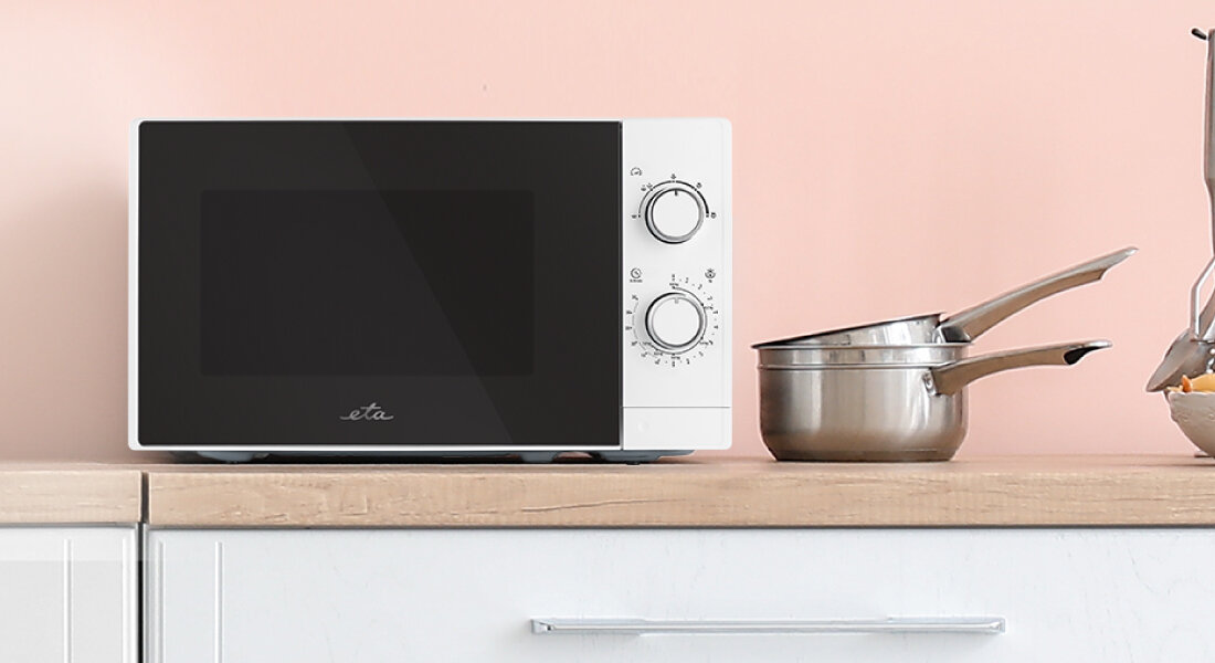 Kuchenka mikrofalowa ETA Marvello ETA421090000 Zdjęcie przedstawia białą kuchenkę mikrofalową ETA ustawioną na drewnianym blacie w jasnej kuchni, obok metalowych garnków. Minimalistyczne tło podkreśla prostą formę urządzenia i kontrast czarnego frontu z jasną obudową