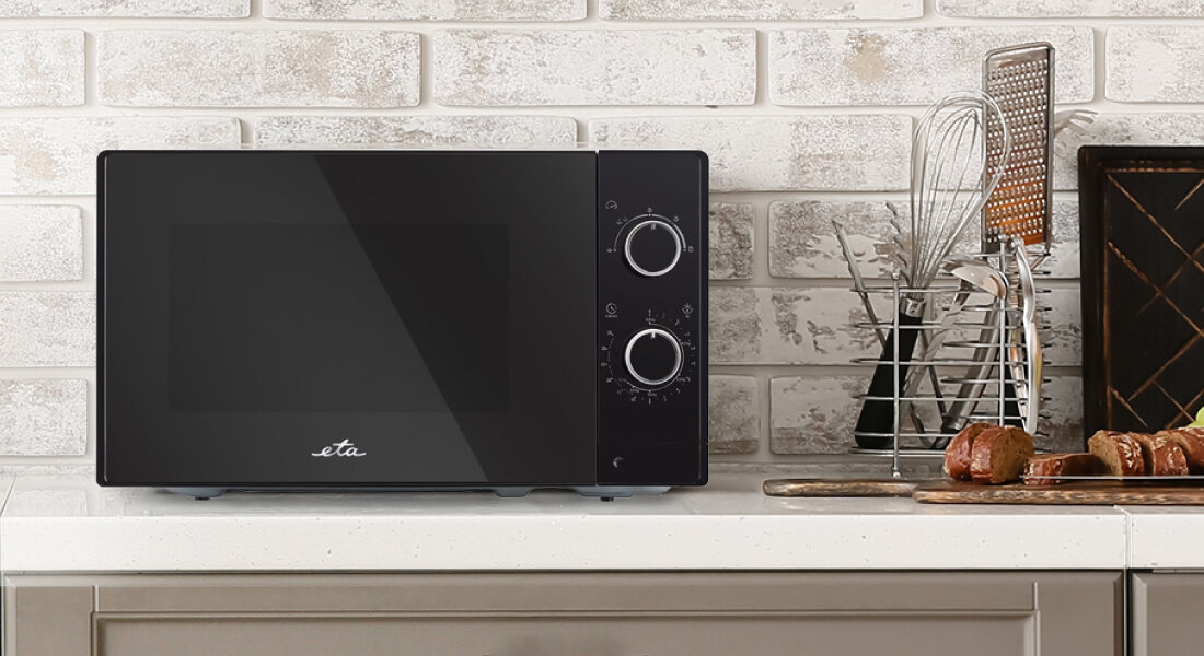 Czarna kuchenka mikrofalowa z dwoma pokrętłami na panelu sterowania, umieszczona na blacie kuchennym. Obok znajdują się akcesoria kuchenne i pieczywo, a na drzwiczkach widoczne jest logo \'eta\'.