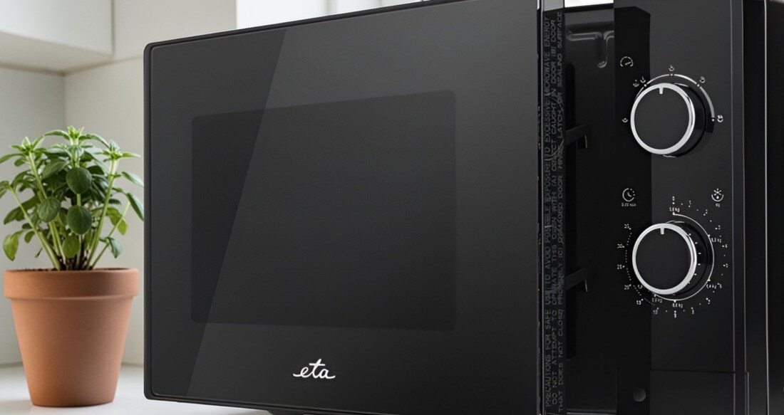 Czarna kuchenka mikrofalowa z logo \'eta\' na drzwiczkach, obok której stoi doniczka z rośliną. Na panelu sterowania znajdują się dwa pokrętła z oznaczeniami liczbowymi.